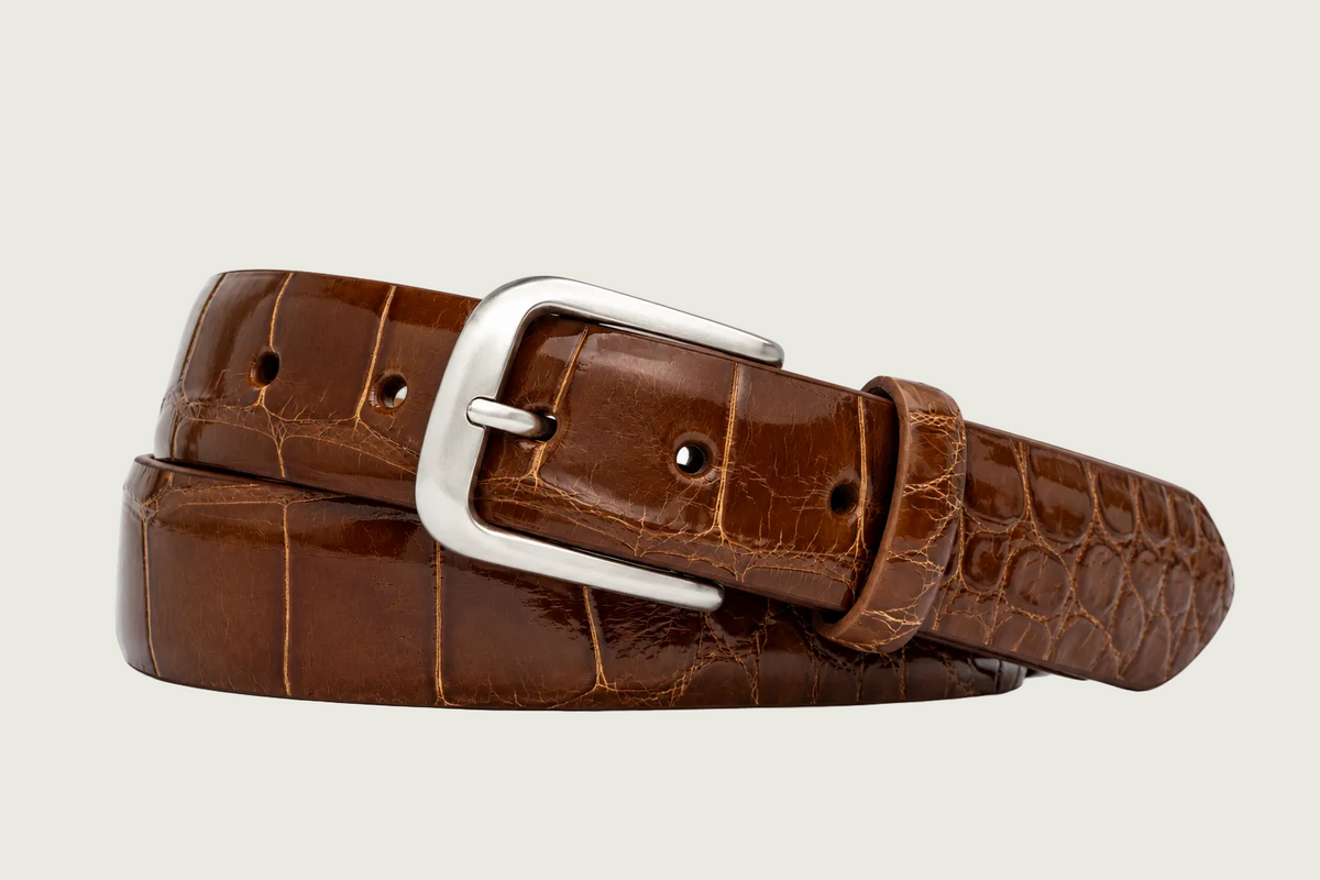 A brown crocodile-embossed leather belt with a polished silver-tone metal buckle, coiled on a white background.