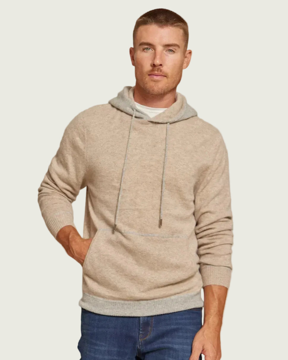 A man wearing a light beige hooded sweater with a contrasting gray hood lining and drawstrings, paired with blue jeans, against a solid gray background.