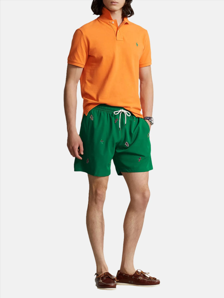 Model is wearing an orange polo shirt and green swim trunks with multi-colored nautical kayak embroidery motifs all over. Trunks have an elastic waistband with a white cord drawstring with metal tips at the end of each drawstring.