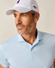 Man wearing a light blue striped polo shirt and white cap with logos on a beige background