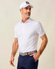 Man wearing a white patterned polo shirt and navy pants with a cap on a plain background